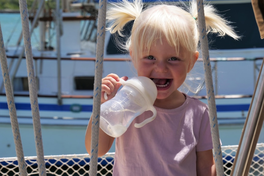 One year old girl with blonde pig tails, smiling with a sippy cup in her mouth, with background of a boat. 
