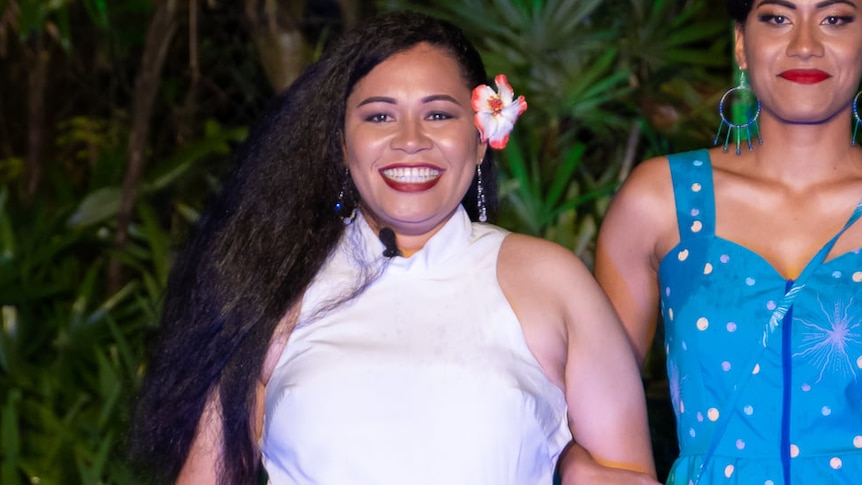 A woman wearing a white top with long dark curly hair and a flower above her ear smiles excitedly at night amid palm trees