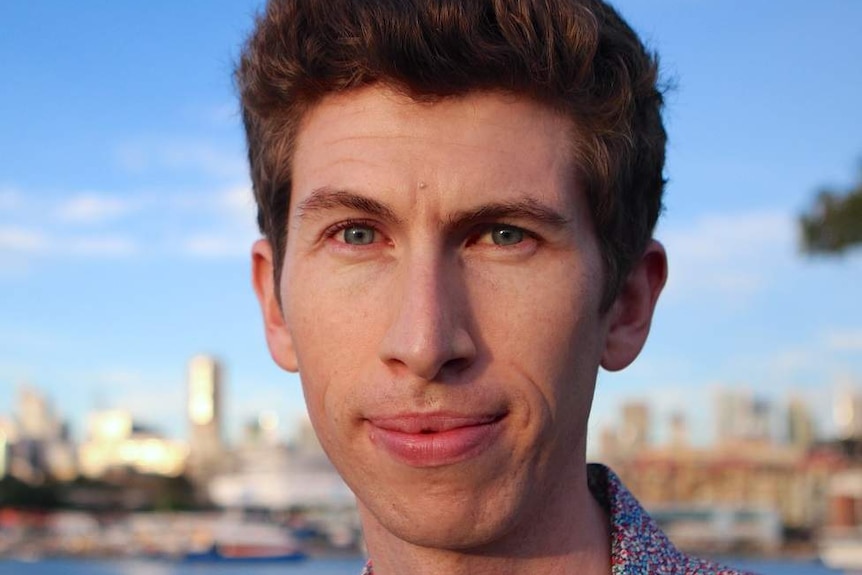 Benjamin Veness stands in front of a bay of water on a sunny day.