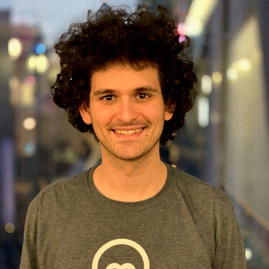 A young man with curly dark hair smiling in front of a window 