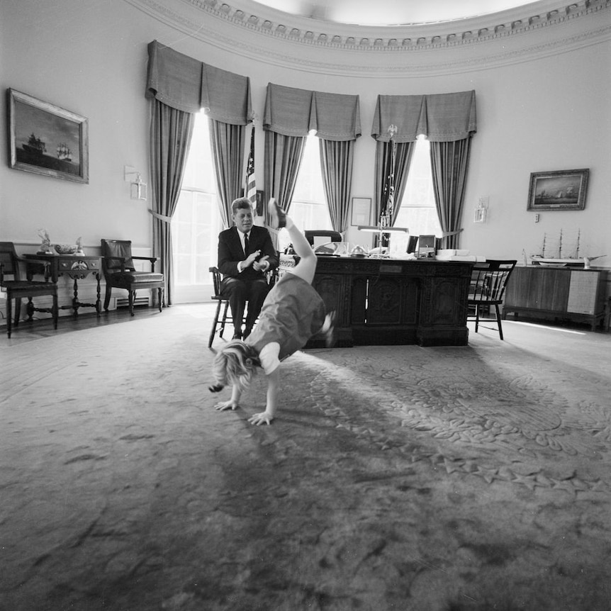A black and white photo of little Caroline Kennedy cartwheeling in the oval office while JFK applauds