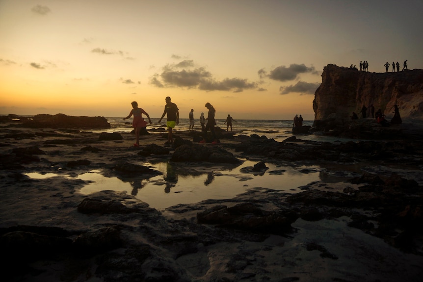 Families are silhouetted against a yellow sunset as they walk between rock pools on a sandy beach.