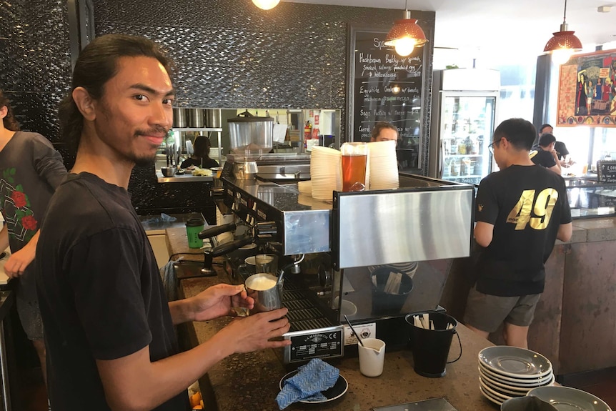 A barista smiles at the camera while making a coffee, with a customer at a counter nearby.
