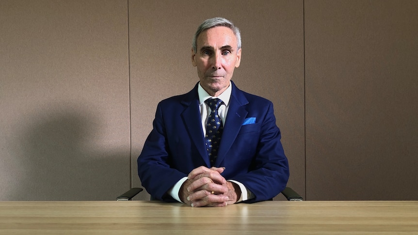 a man with silver hair wearing a dark navy suit sitting at a desk with his hands linked