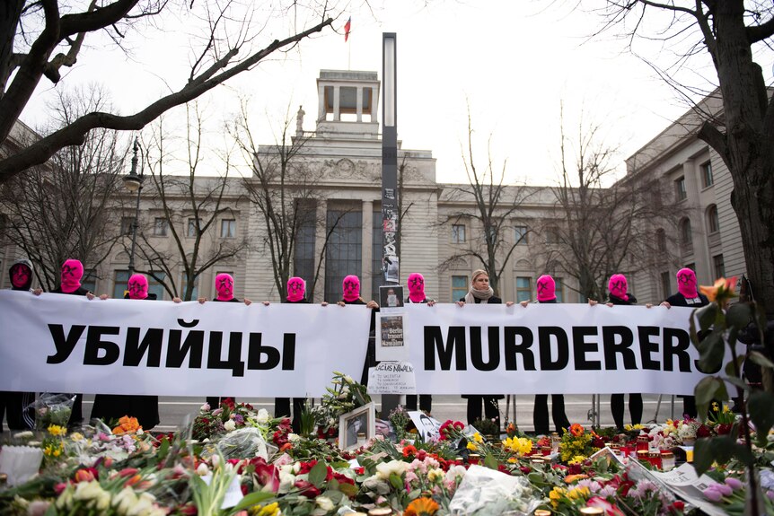 A line of people, most wearing pink masks, hold up signs in Russian and English saying "MURDERERS".