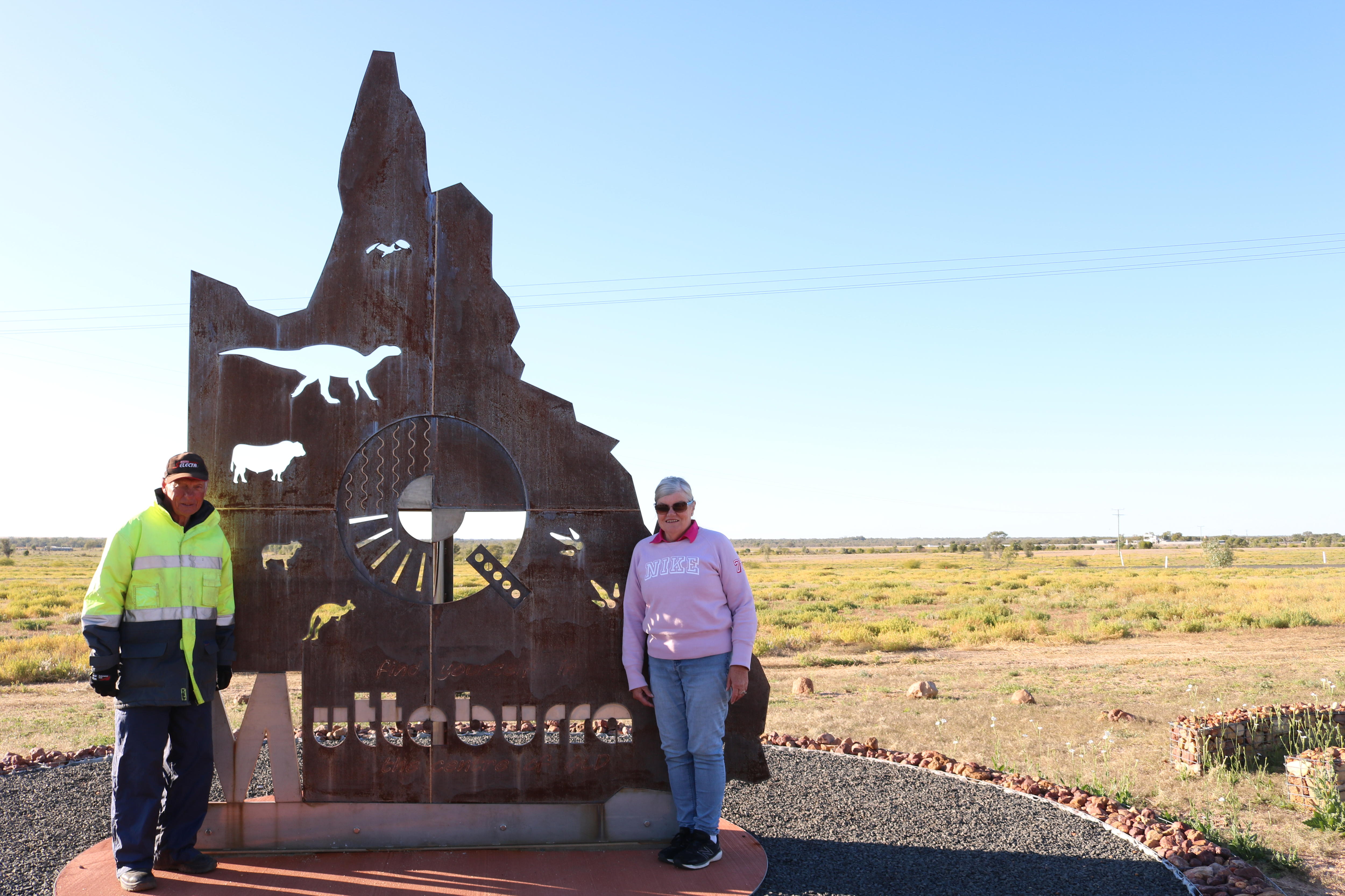 A middle aged man and woman stand in front of a iron cut out reading 'Muttaburra' 