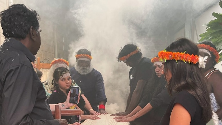 A group of people standing with their hands on a wooden coffin, with smoke in the background, as part of a funeral ceremony. 