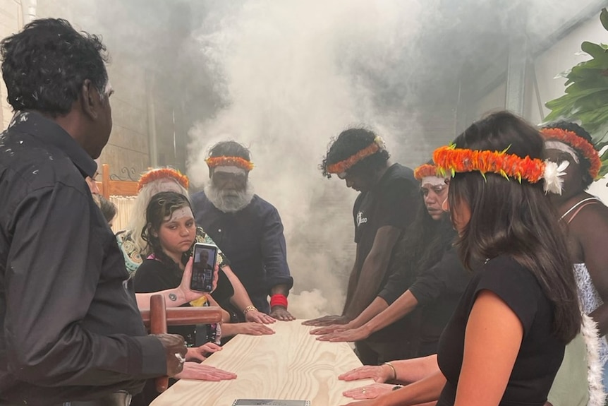 A group of people standing with their hands on a wooden coffin, with smoke in the background, as part of a funeral ceremony. 