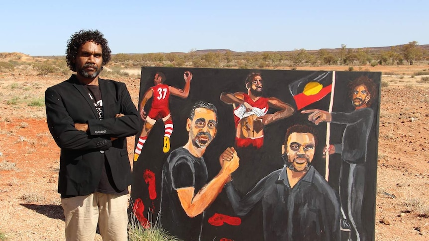 The artist beside his painting - a self-portrait with Adam Goodes - standing outdoors, with landscape behind.