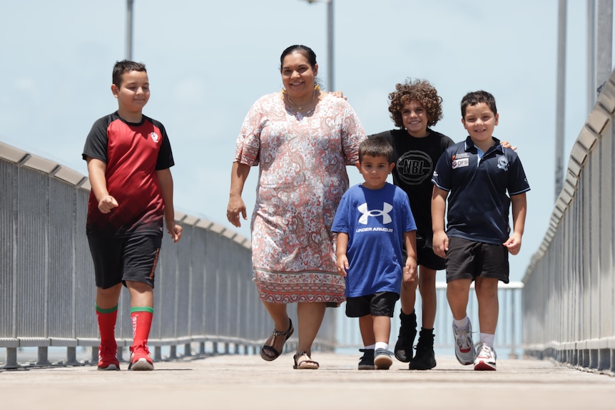 A woman walks along a jetty with 4 boys. 