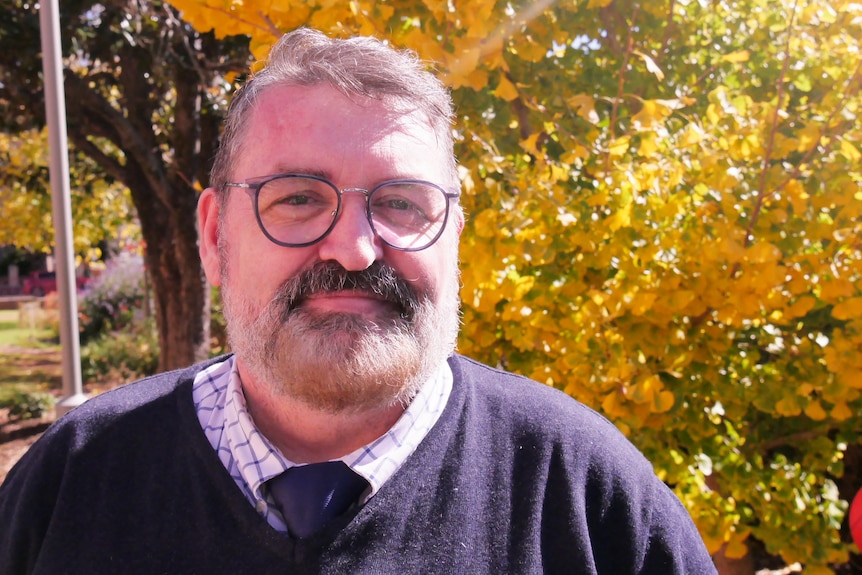 A close-up of a heavy set, grey bearded man in glasses standing in front of a tree with yellow blooms