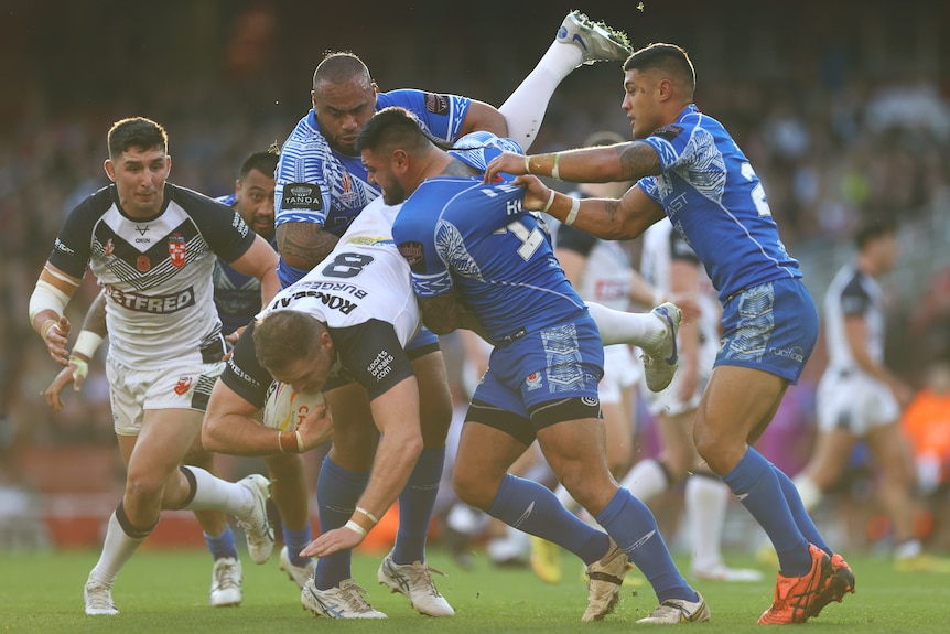 An England rugby league player holds his hand out toward the ground as he is lifted in a tackle by two Samoan forwards.