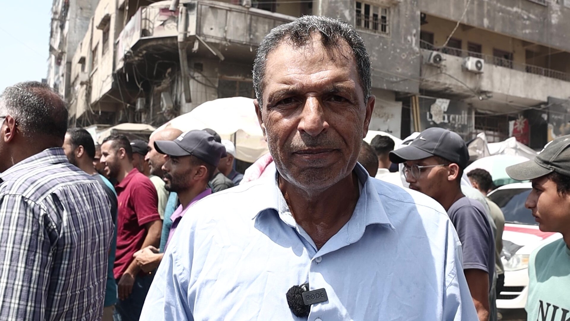A man in a white shirt, standing in front of a crowd and a damaged building.