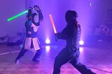 in a dark room two people fight with lightsabers and have protective gear on