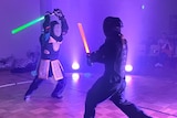 in a dark room two people fight with lightsabers and have protective gear on