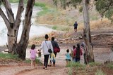 A A group of adults and children walk toward a riverbank, surrounded by gum trees.