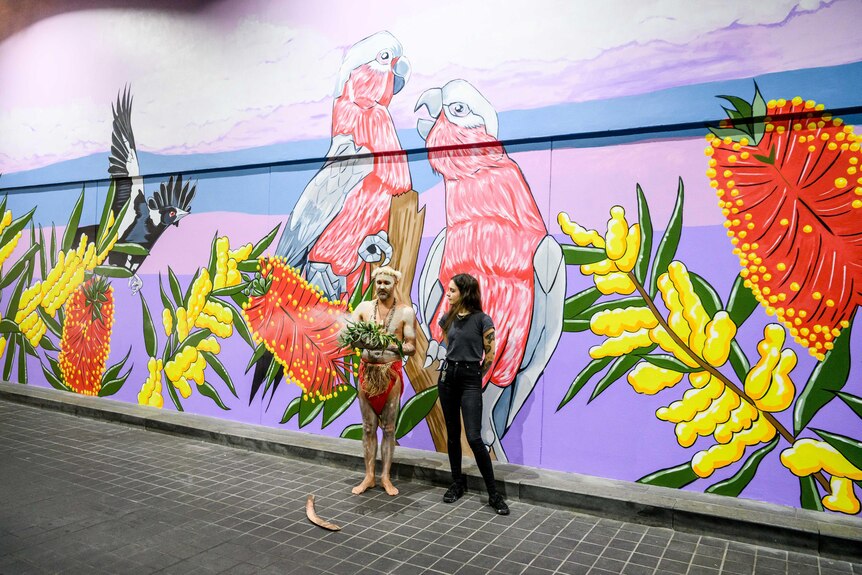 Noni stands with long dark hair next to an Indigenous man in front of a beautiful mural of galahs and native flowers