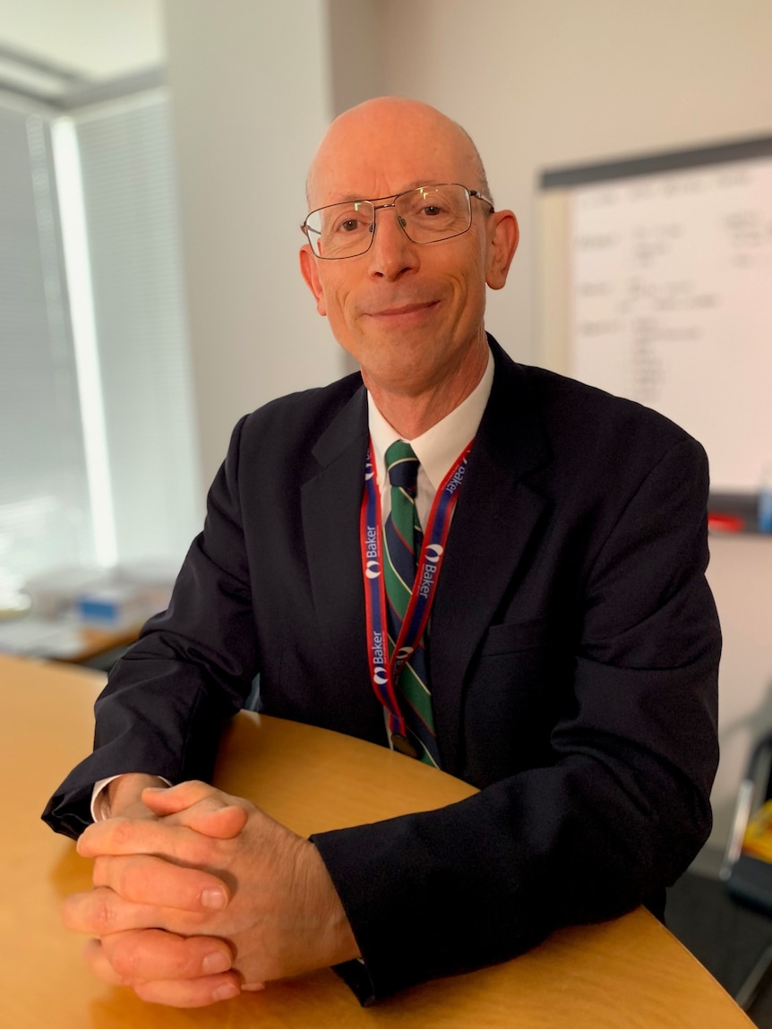 A smiling man wearing spectacles sits at a desk with his hands clasped.