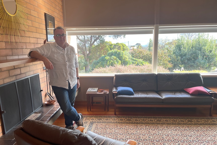 Man leans against fire place mantel piece with windows behind