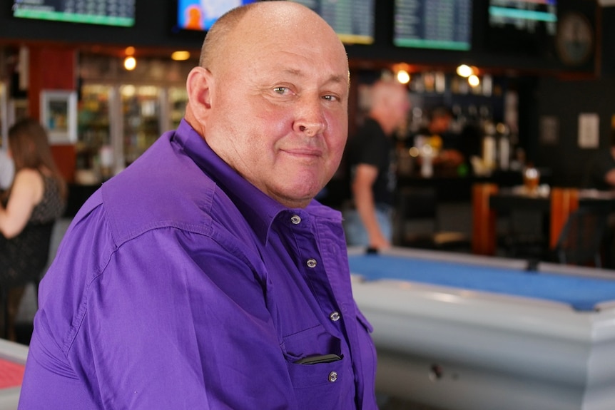 A balding man wearing a purple shirt sits in a pub.