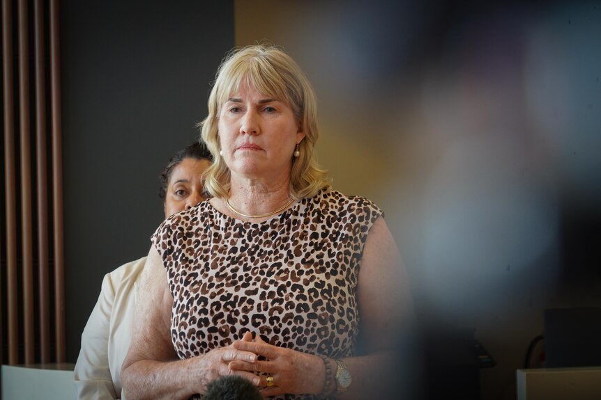 A blonde woman in a leopard-print dress stands with her hands clasped, looking solemn.