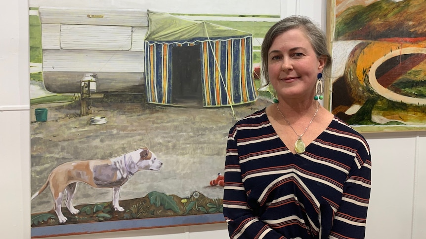 A woman stands in front of a painting of a caravan and tent.