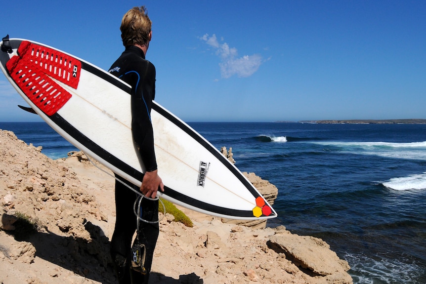 A surfer in a wetsuit holding a surfboard standing on a cliff overlooking the ocean.