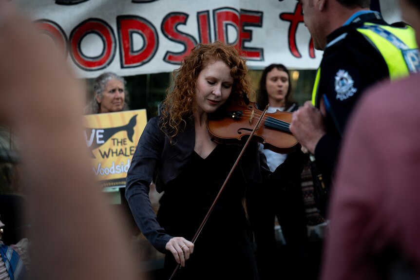 Police stand in front of a woman playing the violin at a protest, banners behind, people holding placards.