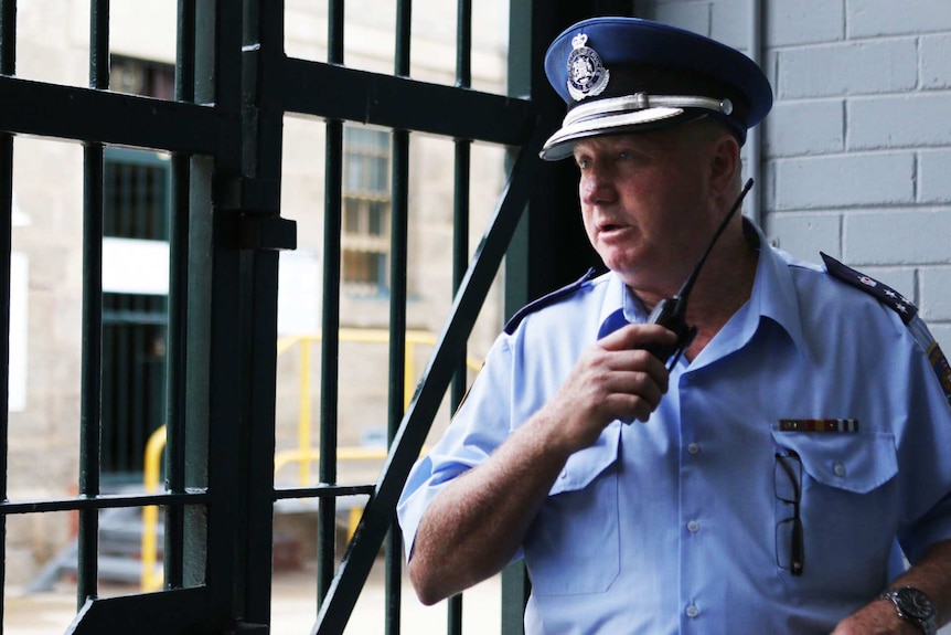 Governor John O'Shea talks on a walkie talkie in front of a heavy, metal fence.