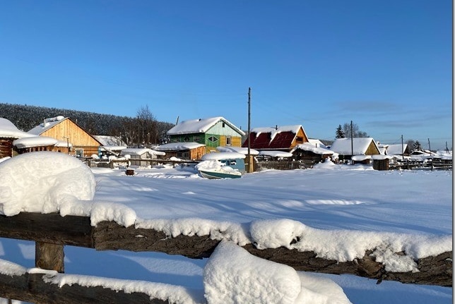 Wooden houses in a village, covered in snow, with a snow covered fence in the foreground.