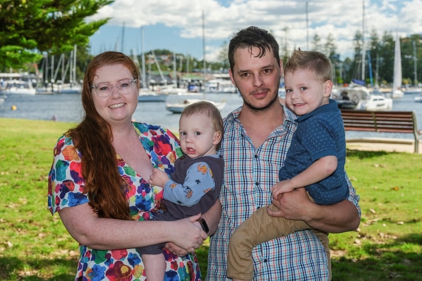 A woman holding a baby stands next to a man holding a four-year-old, they are standing on grass in front of a lake with boats