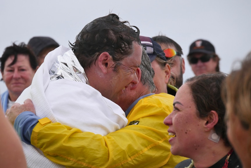 A man wrapped in a white blanket hugs an older man in a yellow jacket. 