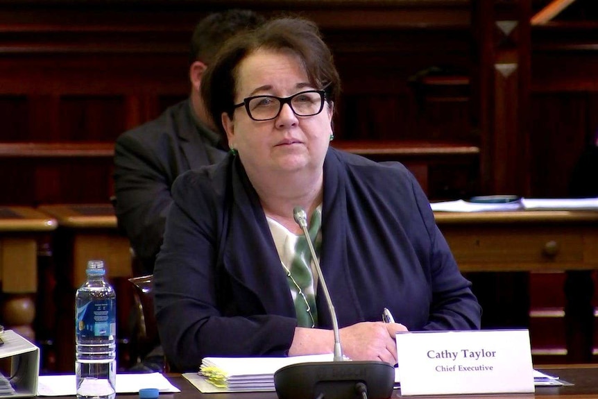 A woman with glasses sitting at a desk in a wood-panelled chamber