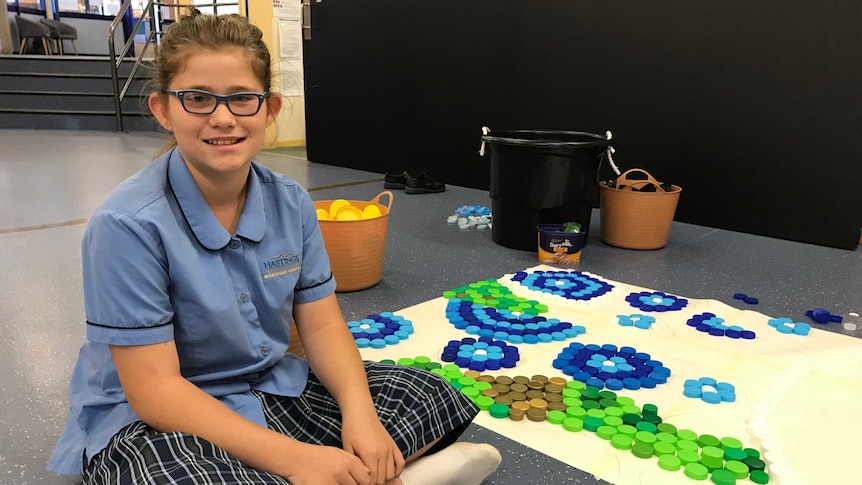 A girl sits with an artwork made of drink bottle lids.