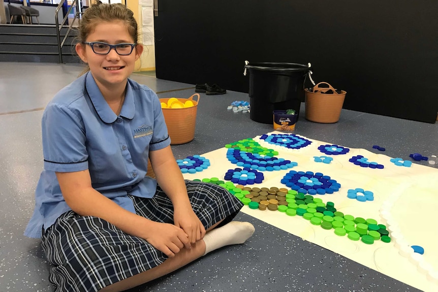 A girl sits with an artwork made of drink bottle lids.