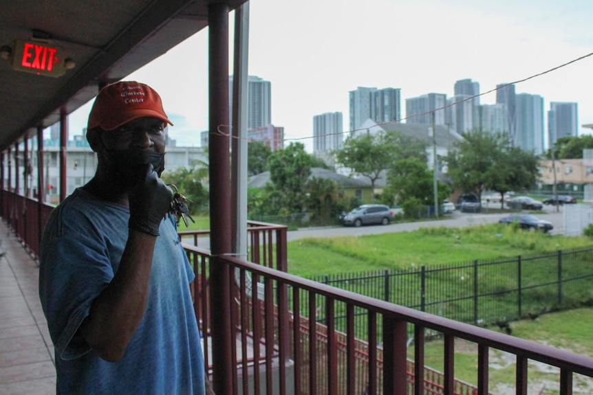 A man in a blue shirt stands on a balcony with several tall skyrises in the background