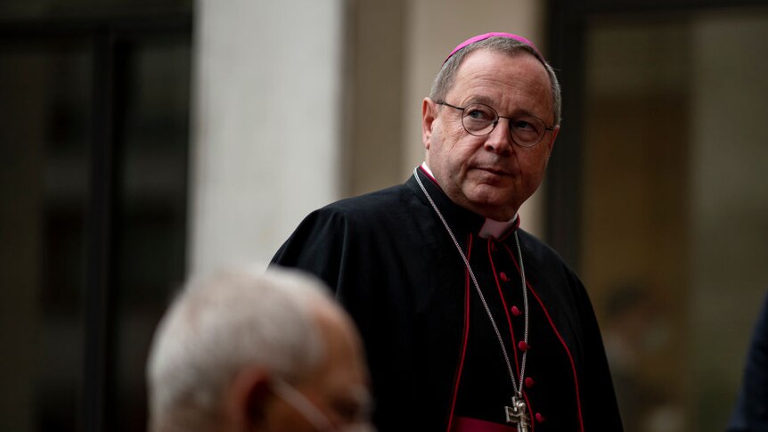 candid shot of Bishop Georg Bätzing in black priest cloak with red lining, pink priest cap and large silver cross necklace
