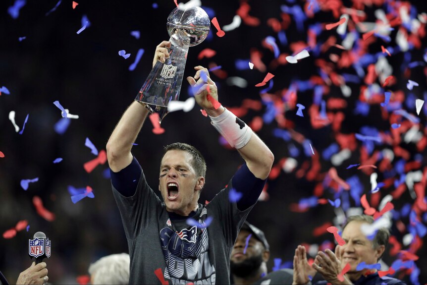A white man with short brown hairs yells in triumph as he holds a silver trophy aloft while red, white and blue streamers fall.