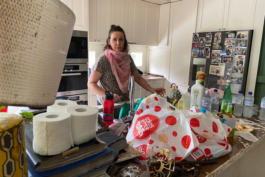 A woman stands in her kitchen with belongings strewn on her bench.