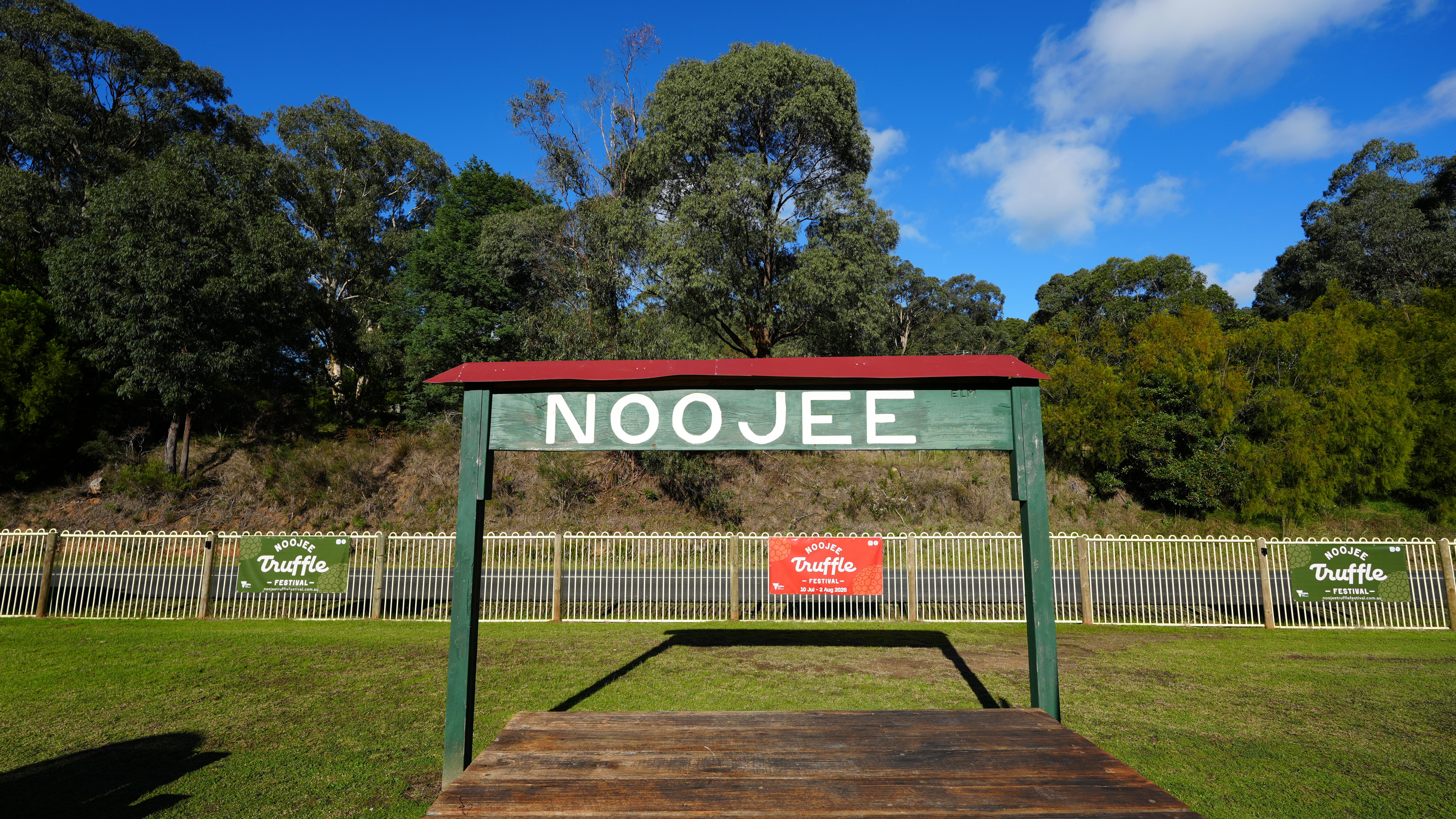 Image of a rail station sign spelling Noojee, with green grass and a blue sky. 