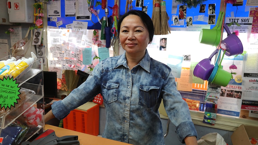 A woman stands behind grocery store counter in front of colourful and cluttered wall.