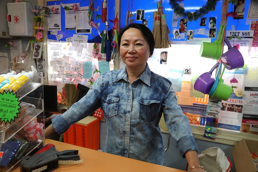 A woman stands behind grocery store counter in front of colourful and cluttered wall.