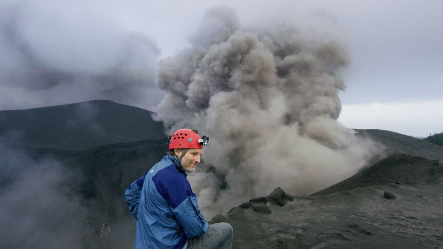 Explorer Carsten Peters wears a red helmet and blue jacket and stands close to the grey smoky rim of a volcano.