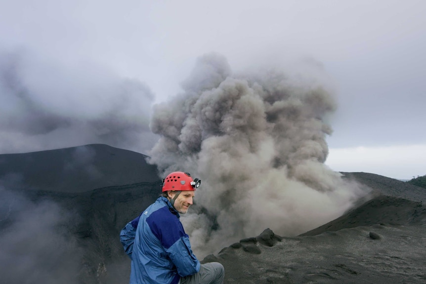 Explorer Carsten Peters wears a red helmet and blue jacket and stands close to the grey smoky rim of a volcano.