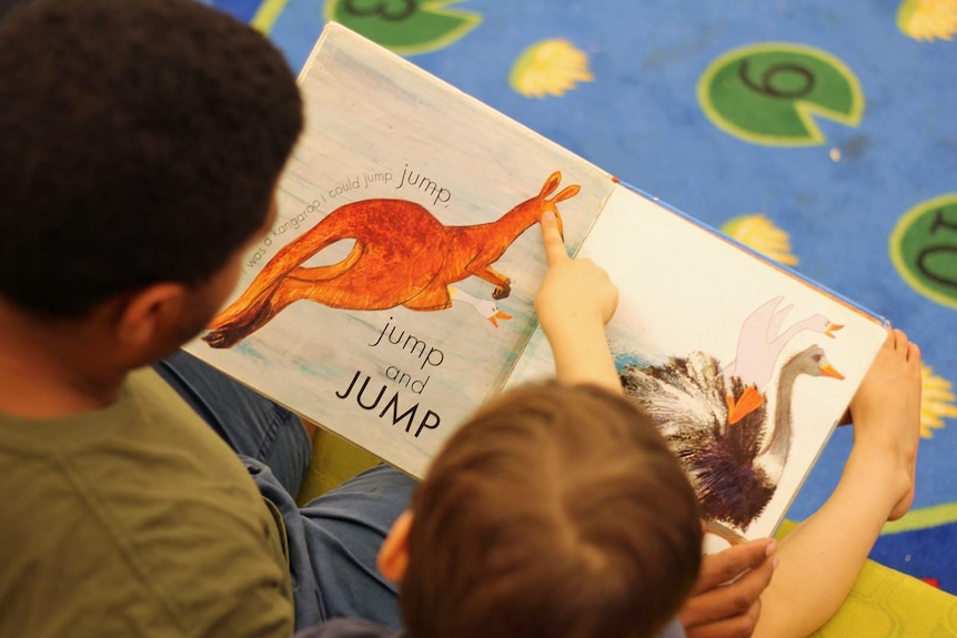 A man and male child sit on a couch reading a book together, you can't see their faces but the child is pointing at the pictures