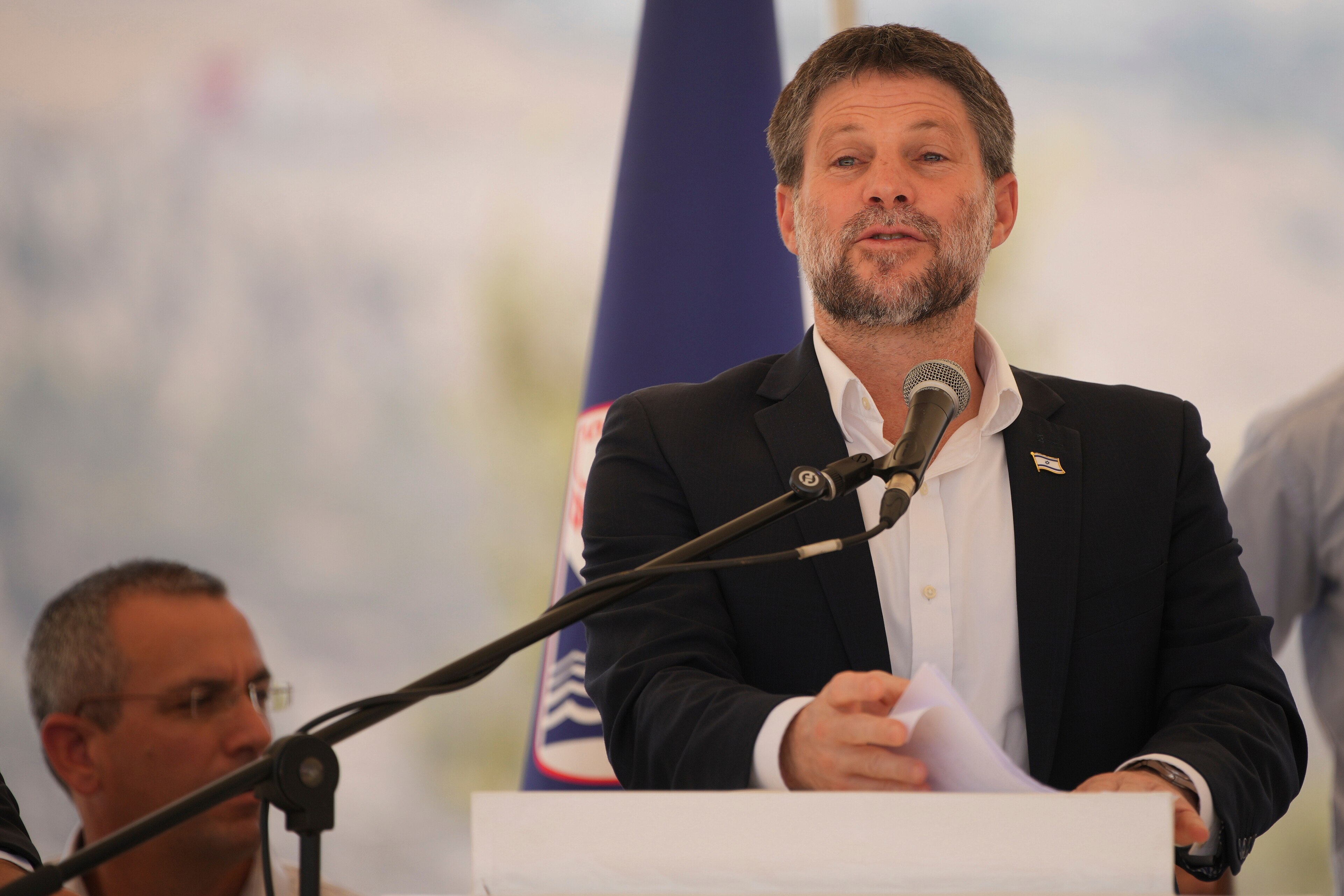 A bearded man in a suit speaks confidently in front of a blue Israeli flag.