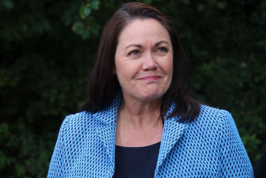 Liza Harvey wearing a light blue jacket, standing in front of dark green shrubs.