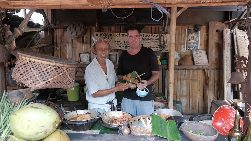 Two men stand under a bamboo hut cooking food