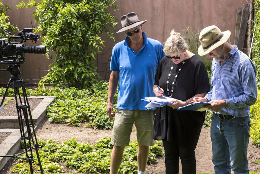 Lomas and two men looking at clipboards with camera standing next to them on a tripod in a garden.