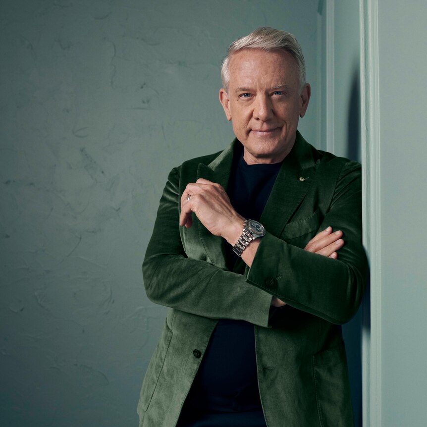 Simon Burke, wearing a green jacket, leans against a wall with his arms folded and smiles at the camera.